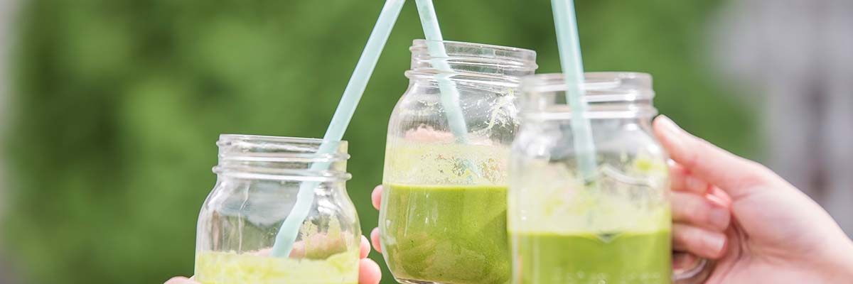 3 green smoothies in glass mason jars with handles and straws being cheered