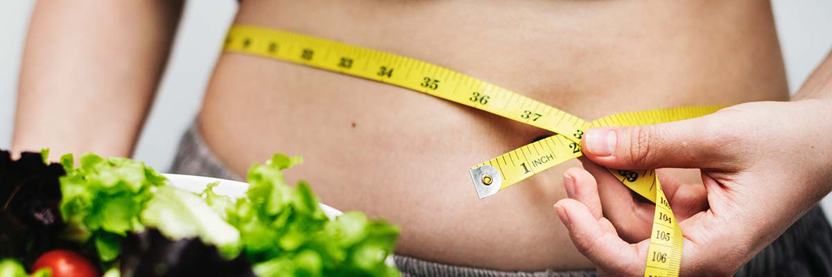 close up of woman measuring her waist with a fabric tape measure while holding a salad