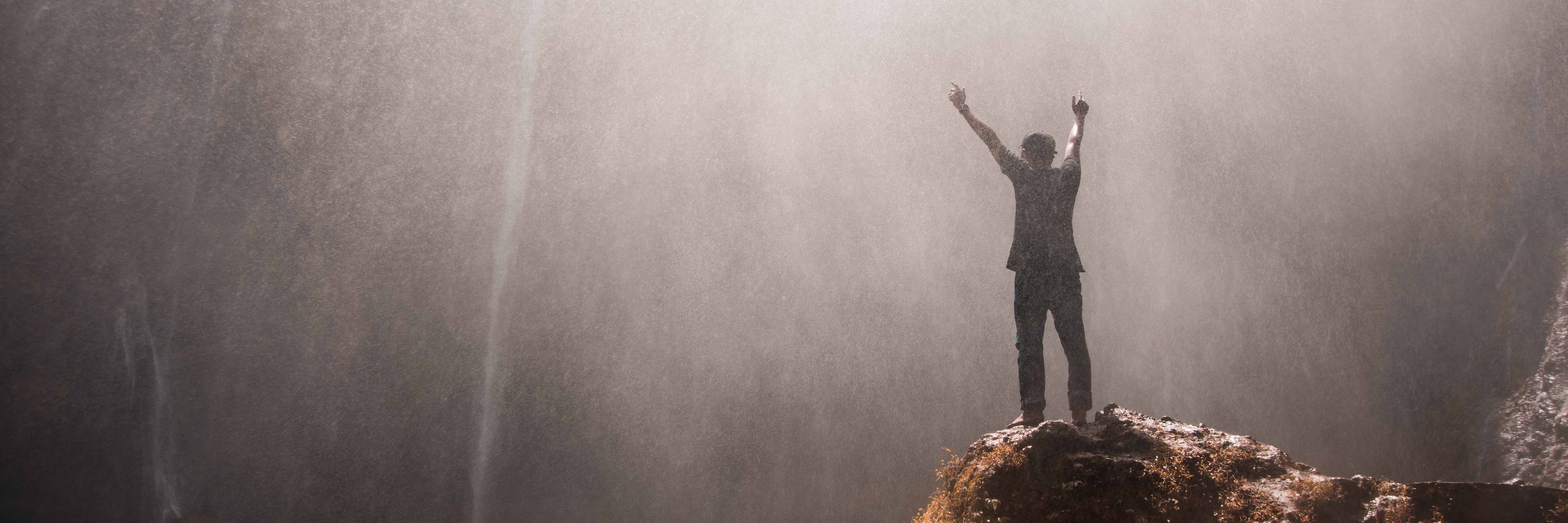 man stading in front of waterfall with arms raised above head